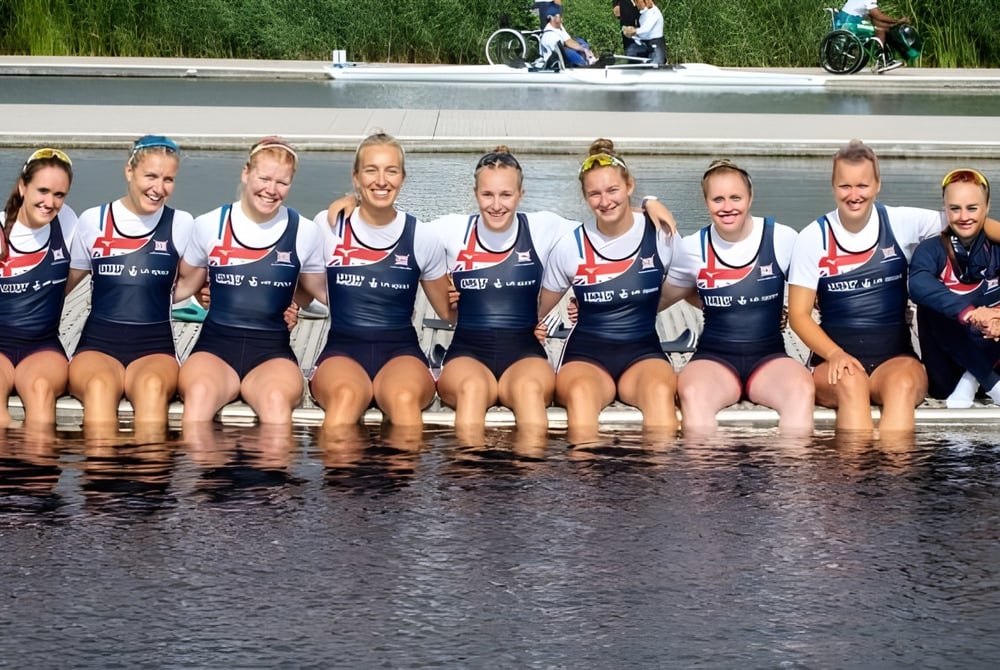 Eine Gruppe von Athletinnen der Durham School sitzt im Wasser mit Gras und Radfahrern im Hintergrund.