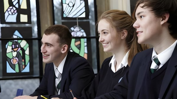 Drei Schülerinnen und Schüler der Durham School stehen in Schuluniform vor einem Buntglasfenster und lächeln.