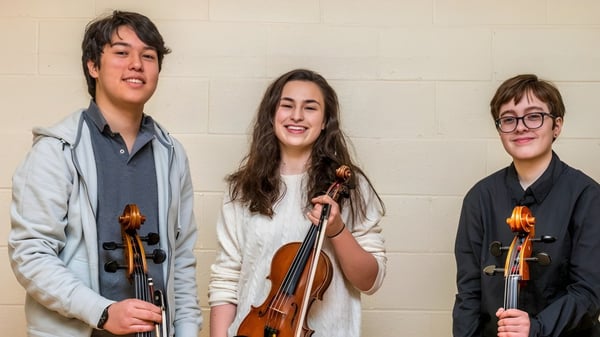 Drei Schüler der Eagle School halten Musikinstrumente und stehen vor einer schlichten Wand.