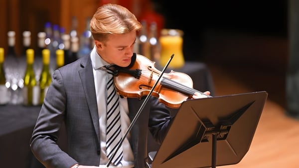 Ein junger Mann spielt in einem Anzug Violine auf der Bühne der Earlscliffe School mit Weinflaschen im Hintergrund.