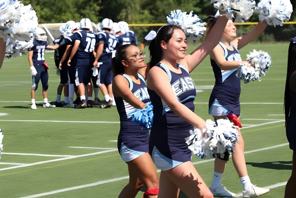 Cheerleader der East Catholic High School führen eine Performance auf dem Footballfeld mit Spielern im Hintergrund aus.