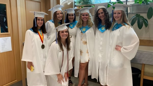 Eine Gruppe von Absolventinnen der East Catholic High School in weißen Talaren mit Medaillen steht zusammen im Schulgebäude.