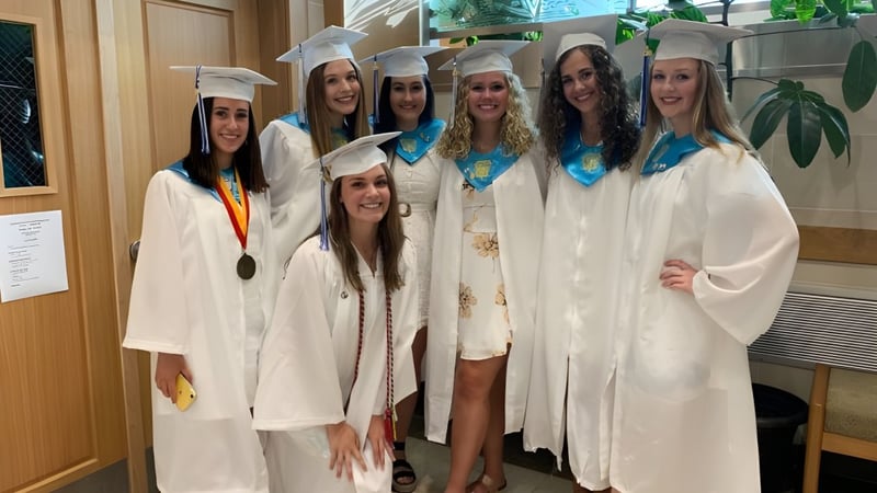 Eine Gruppe von Absolventinnen der East Catholic High School in weißen Talaren mit Medaillen steht zusammen im Schulgebäude.