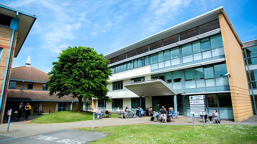 Das moderne mehrstöckige Gebäude des East Sussex College Lewes ist von einer Rasenfläche und einem großen Baum umgeben, während Schülerinnen und Schüler das Gebäude betreten und verlassen.