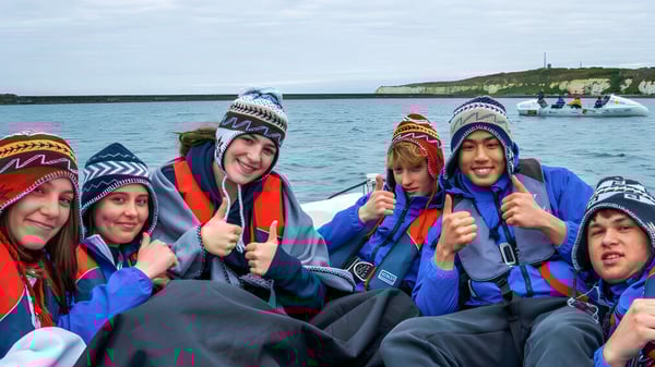 Eine Gruppe von Schülern des Eastbourne College trägt bunte Kapuzenjacken und sitzt gemeinsam auf einem Boot vor einer Klippenlandschaft.