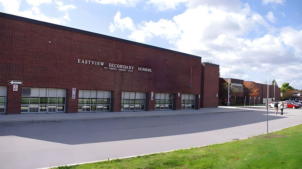 Das große Backsteingebäude der Eastview Secondary School vor einem bewölkten Himmel mit gepflastertem Bereich und Rasenfläche.