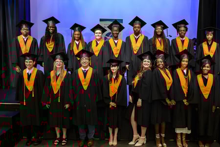Absolventen der École secondaire publique L'Alternative stehen in schwarzen und orangen Talaren auf einer Bühne vor blauem Hintergrund.