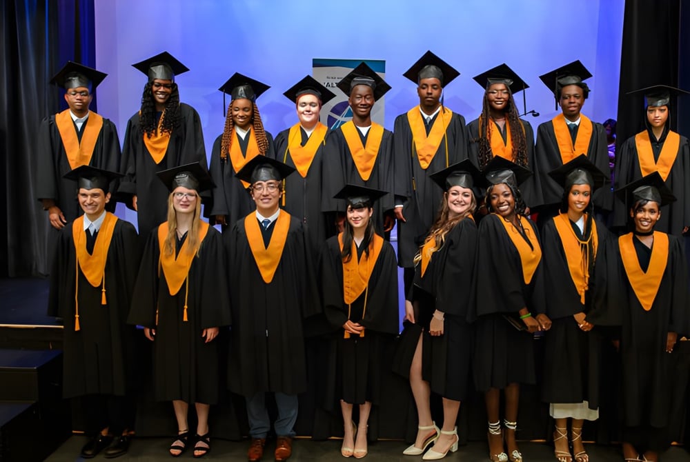 Absolventen der École secondaire publique L'Alternative stehen in schwarzen und orangen Talaren auf einer Bühne vor blauem Hintergrund.