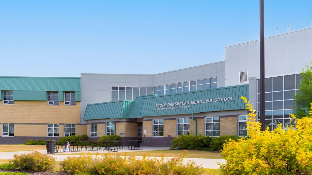 Das moderne Schulgebäude der École Dansereau Meadows School mit grünem Dach und gelben Blumen im Vordergrund unter blauem Himmel.