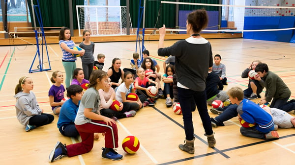 Eine Gruppe Schüler sitzt in der Turnhalle der École des Deux-Rives und hört aufmerksam einer erwachsenen Lehrkraft zu.