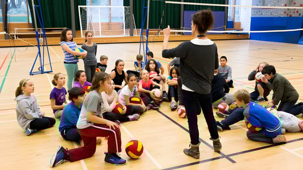 Eine Gruppe Schüler sitzt in der Turnhalle der École des Deux-Rives und hört aufmerksam einer erwachsenen Lehrkraft zu.