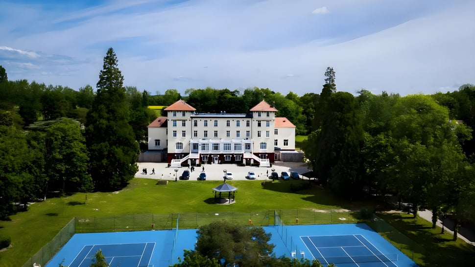 Das Gebäude der Ecole des roches steht mit rotem Ziegeldach umgeben von Grünflächen und blickt auf einen Hof mit Tennisplätzen und einem Brunnen.