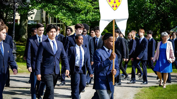 Schüler der Ecole des roches in formeller Kleidung tragen eine Flagge und gehen durch einen Park mit viel Grün.