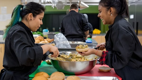 Zwei Schülerinnen der École des Sept Sommets servieren Essen aus einer großen Schüssel während andere Menschen im Hintergrund sind.