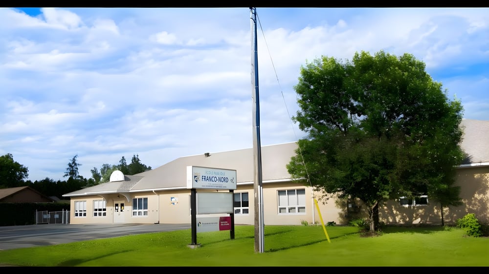 Das moderne beige Gebäude der École Franco-Nord steht vor einem großen Baum unter blauem Himmel mit vereinzelten Wolken.