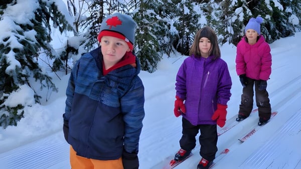Drei Personen in warmer Winterkleidung stehen auf einem verschneiten Weg umgeben von schneebedeckten Kiefern auf dem Gelände der École Franco-Nord.