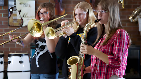 Drei Schülerinnen der École J.E. Lapointe School spielen gemeinsam Blechblasinstrumente in einem Raum mit Backsteinwänden.