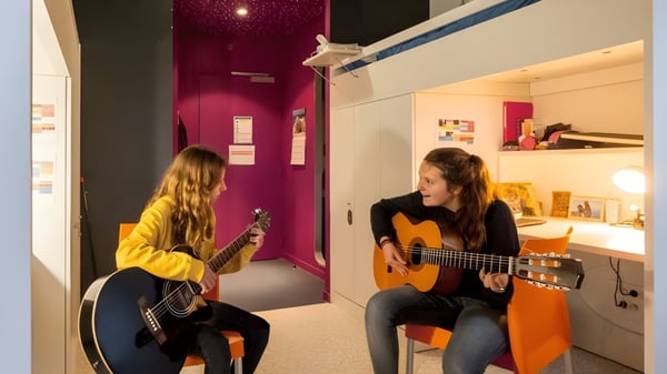 Zwei Schülerinnen spielen auf dem Campus der École Jeannine Manuel akustische Gitarren vor einer pinken Wand.