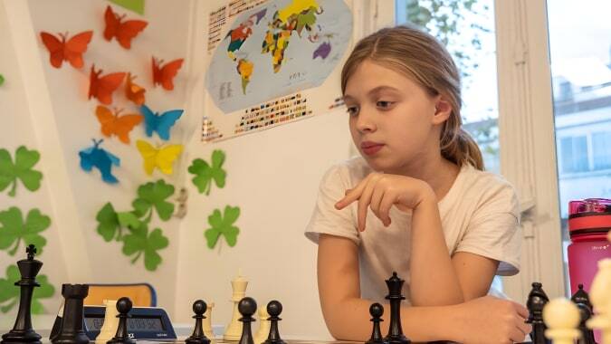 Ein Mädchen spielt konzentriert Schach auf dem Campus der École Jeannine Manuel vor einer Weltkarte und bunten Dekorationen.