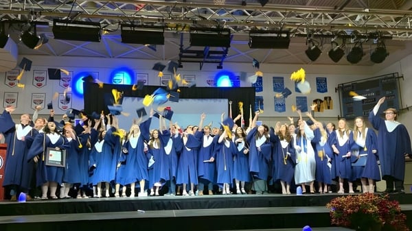 Absolventen der École secondaire publique Le Sommet stehen auf der Bühne in ihren blauen Abschlusstagungen mit Bühnenbeleuchtung im Hintergrund.