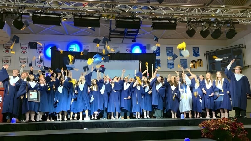 Absolventen der École secondaire publique Le Sommet stehen auf der Bühne in ihren blauen Abschlusstagungen mit Bühnenbeleuchtung im Hintergrund.