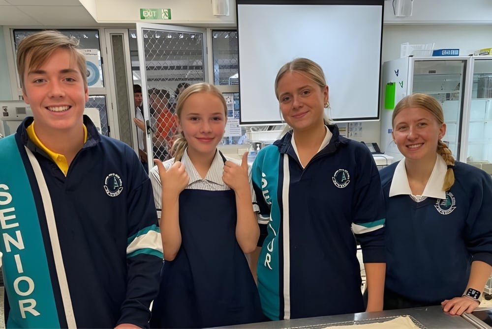 Vier Schülerinnen und Schüler der Elanora State High School in Schuluniform lächeln in einer schulischen Umgebung in die Kamera.