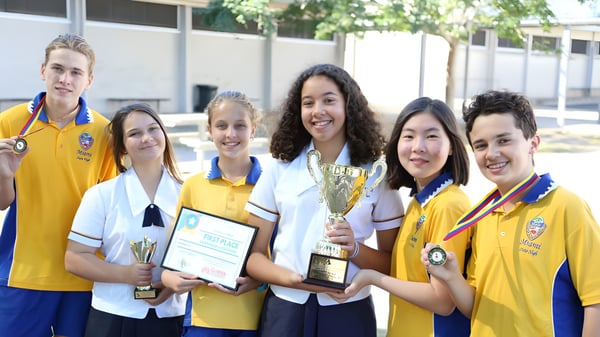Eine Gruppe von Schülerinnen und Schülern der Elanora State High School hält Trophäen und Zertifikate vor dem Schulgebäude mit Bäumen im Hintergrund.