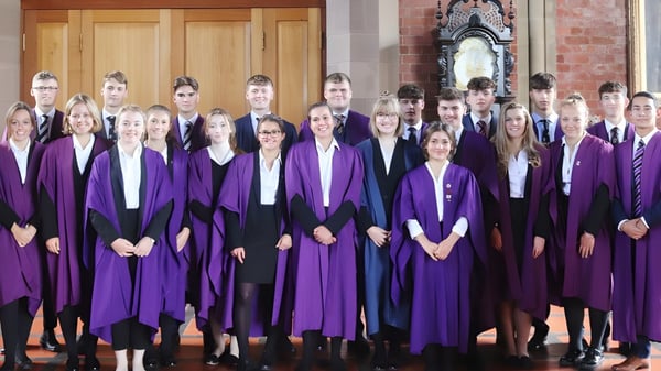 Eine Gruppe von Absolventen steht in lila Talaren vor einem Backsteingebäude mit einem verzierten Torbogen auf dem Campus des Ellesmere College.