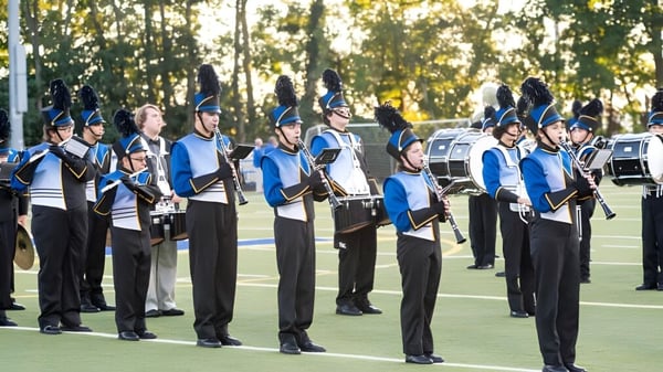 Mitglieder der Marching Band der Elmwood High School stehen in Formation auf einem Grasfeld vor Bäumen und blauem Himmel.