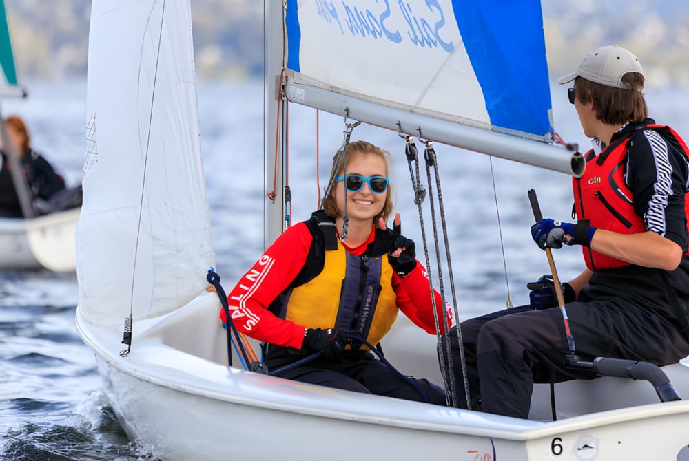 Zwei Personen sitzen in einem kleinen Segelboot auf dem Wasser mit blau-weißem Segel im Hintergrund auf dem Gelände der Embley.