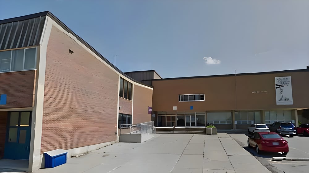 Das moderne Backsteingebäude mit großen Fenstern vom Emery Collegiate Institute ist von einem gepflasterten Bereich mit geparkten Autos umgeben.