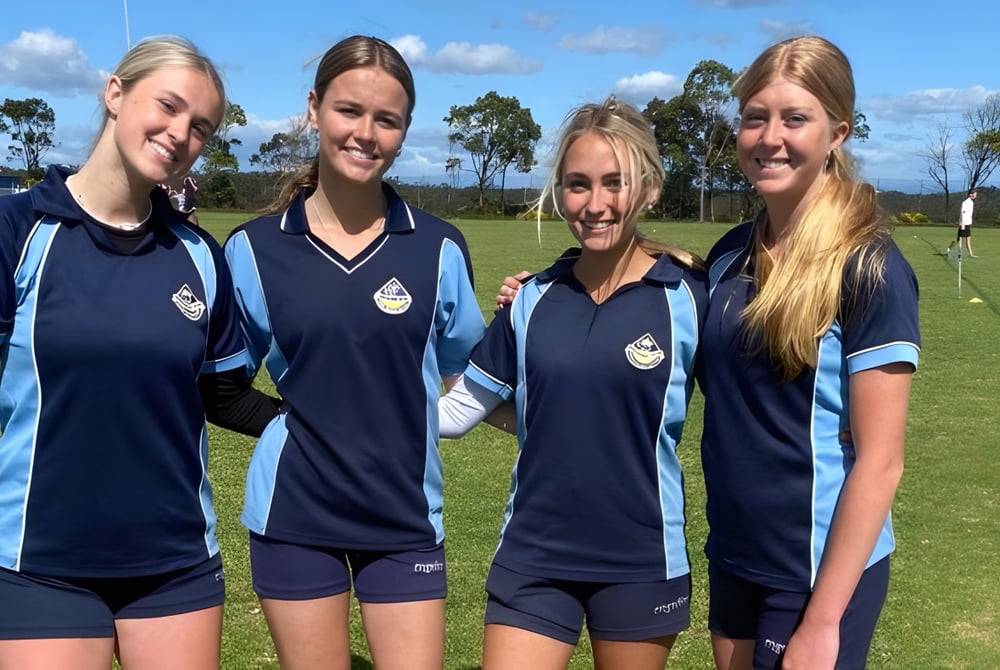 Vier Schülerinnen der Engadine High School stehen zusammen auf einem Sportfeld mit Bäumen und blauem Himmel im Hintergrund.
