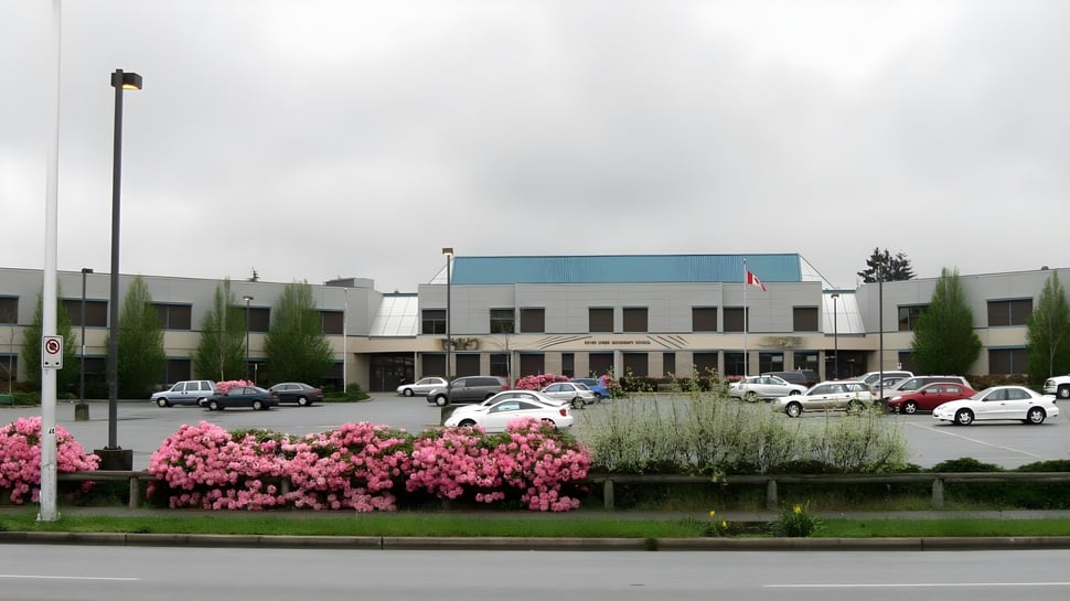 Das Gebäude der Enver Creek Secondary School mit einem Parkplatz im Vordergrund und blühenden rosa Blumen unter bewölktem Himmel.