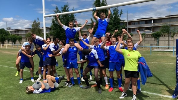 Eine Gruppe von Schülern der Episcopal Sem' De Walbourg feiert gemeinsam auf einem Sportplatz mit Schulgebäuden im Hintergrund.