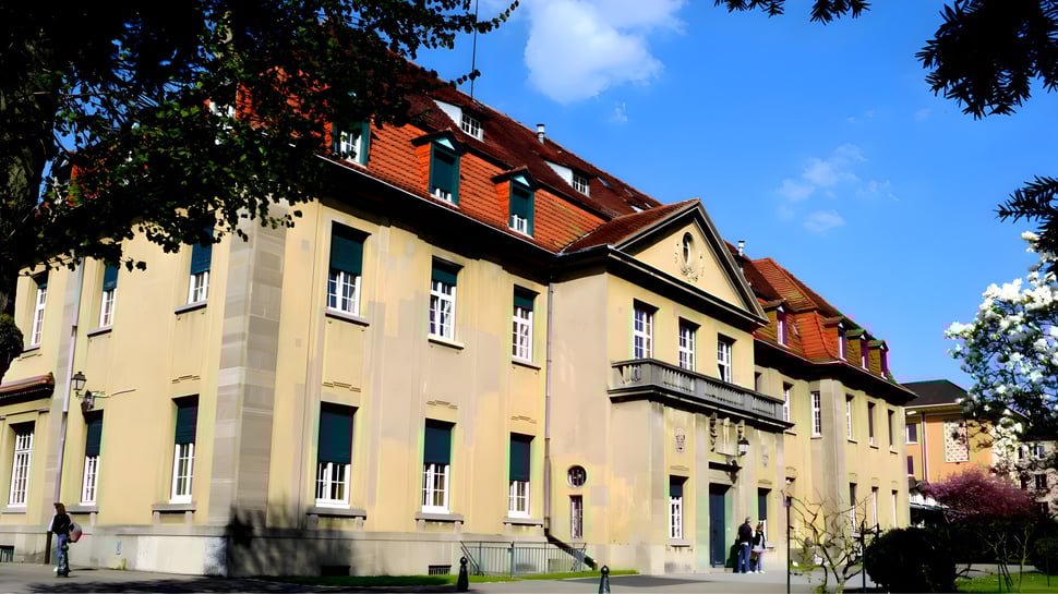 Das mehrstöckige Gebäude der Episcopal Sem' De Walbourg mit rotem Ziegeldach steht umgeben von Grünflächen unter blauem Himmel.