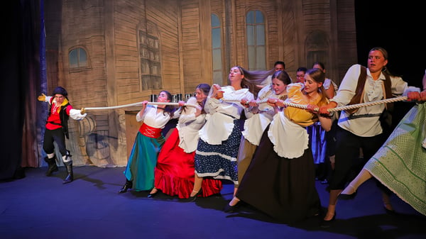 Eine Gruppe von Schülerinnen und Schülern des Epsom College trägt bunte traditionelle Kostüme auf einer Bühne mit Holzverkleidung und Bogenfenstern.
