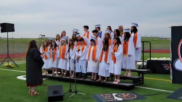 Eine Gruppe von Schülerinnen der Erie High School steht in weißen und orangen Roben auf einer Bühne im Freien vor Lautsprechern.