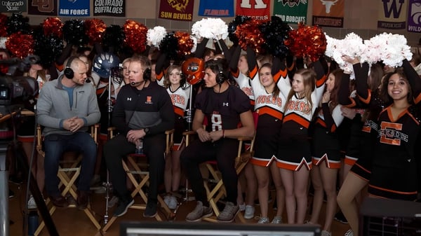 Eine Gruppe Cheerleader in orange-schwarzen Uniformen steht mit Pompons vor den Sport-Bannern der Erie High School.