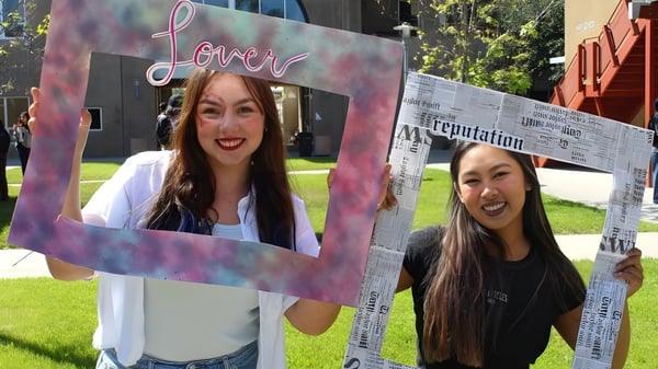 Zwei Schülerinnen der Escondido Charter High School halten einen großen herzförmigen Rahmen auf einer Wiese vor dem Schulgebäude.