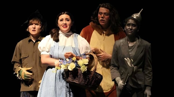 Eine Gruppe von Schülerinnen und Schülern der Esquimalt High School steht in historischen Kostümen mit Blumen und Körben vor dunklem Hintergrund.