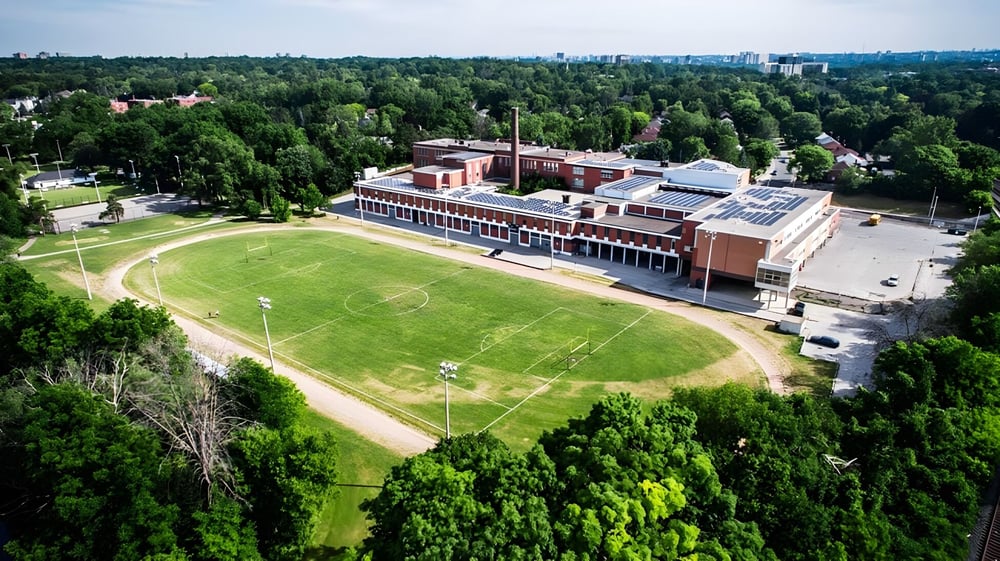 Das große Backsteingebäude des Etobicoke Collegiate Institute steht vor einem grünen Feld mit Wald im Hintergrund.