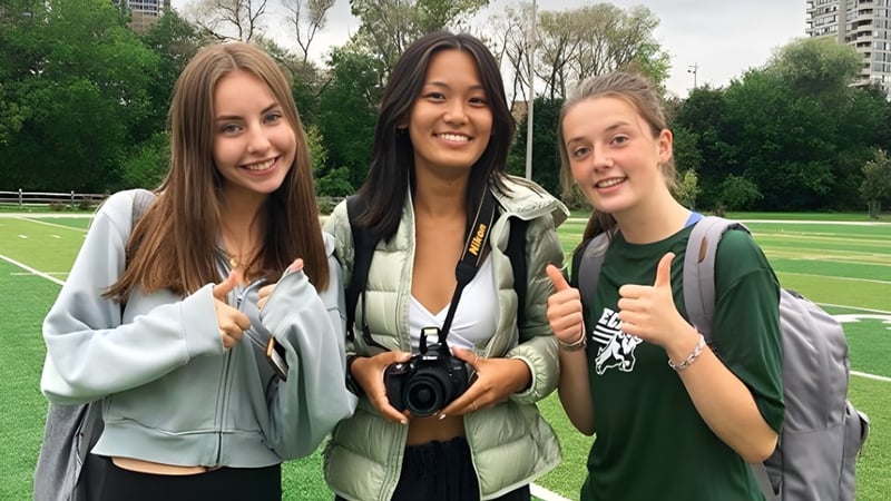 Drei Schülerinnen stehen lächelnd im Grünen auf dem Campus des Etobicoke Collegiate Institute und zeigen jeweils einen Daumen nach oben.