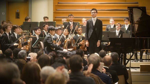 Ein Orchester des Eton College tritt auf der Bühne mit einem Dirigenten und einem Pianisten am Fluegel auf.