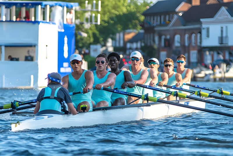 Eine Gruppe von Ruderern in einheitlichen türkisfarbenen Uniformen sitzt im Ruderboot auf dem Wasser vor einer malerischen Kulisse auf dem Campus des Eton College.