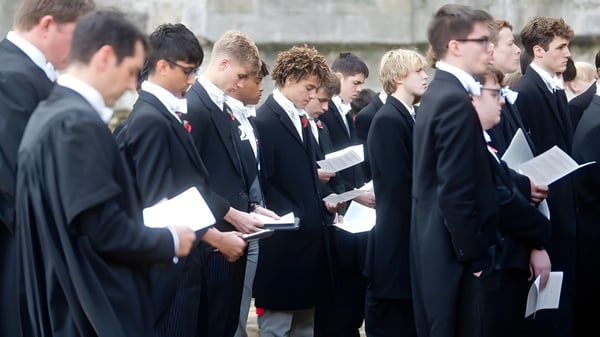 Eine Gruppe von Schülerinnen und Schülern des Eton College steht in formeller Kleidung vor einem Steingebäude und hält Dokumente in der Hand.
