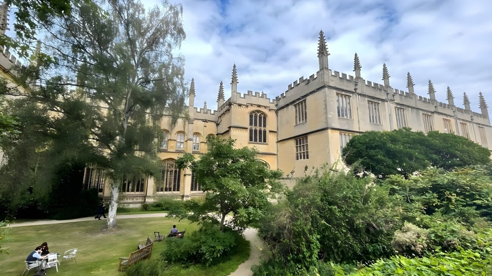 Vor der gotischen Kathedrale auf dem Campus des Exeter College sitzt eine Person auf einer Bank zwischen Bäumen.