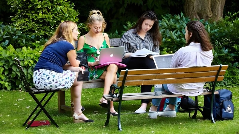 Schülerinnen des Exeter College sitzen auf einer Bank im grünen Außenbereich und lernen gemeinsam mit Laptop und Büchern.