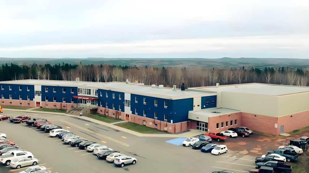 Das große Gebäudekomplex der Exploits Valley High School mit einem Parkplatz im Vordergrund und Wald im Hintergrund.