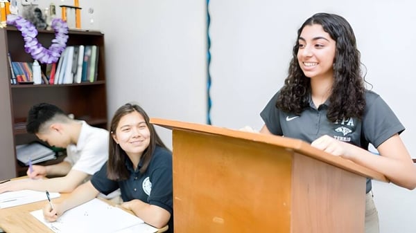 Drei Schülerinnen der Fairmont Prep Academy sind im Klassenzimmer, eine steht am Podium, zwei sitzen an Tischen.