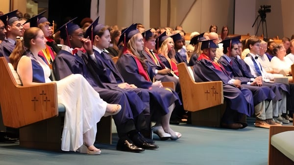 Absolventinnen und Absolventen der Faith Christian School sitzen in Talaren bei einer Abschlussfeier mit Bühne und Publikum.